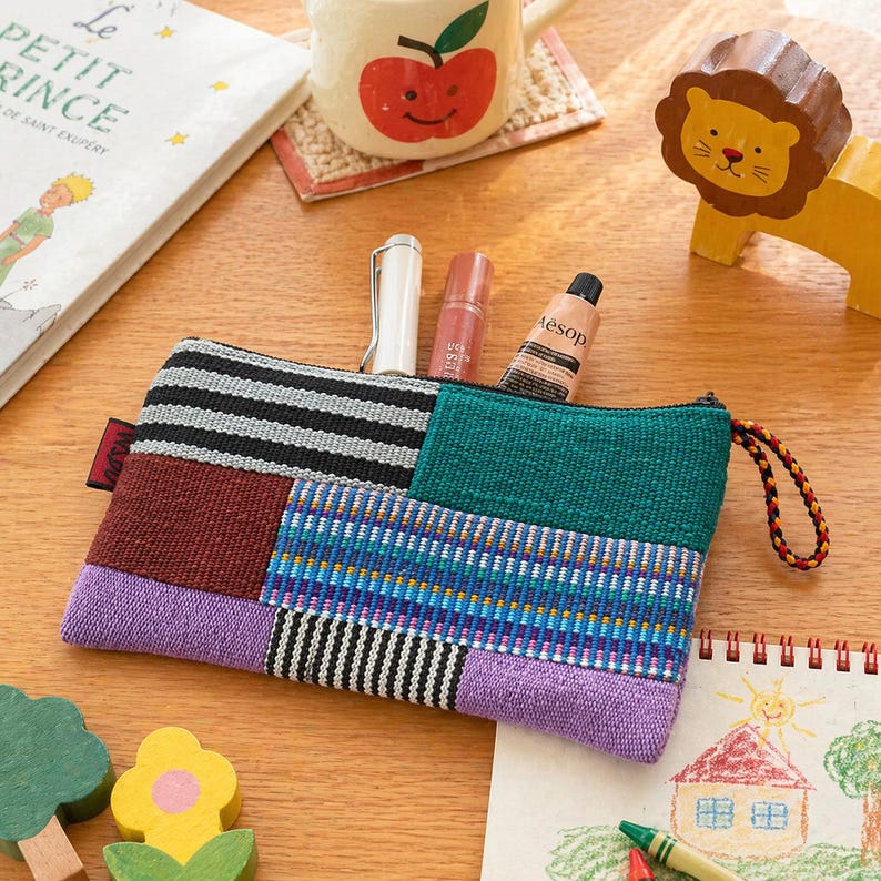 May include: A colourful patchwork zipped pouch with a variety of patterns, including stripes and geometric designs. The pouch contains a pen, lip gloss, and a tube of Aesop hand cream. A children's book and a wooden lion toy are also visible.