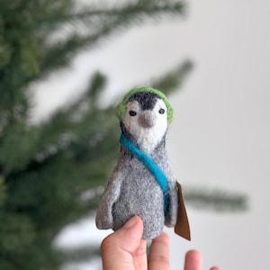 May include: A small grey felt penguin wearing a green felt hat and a blue felt scarf. The penguin is holding a small brown tag.