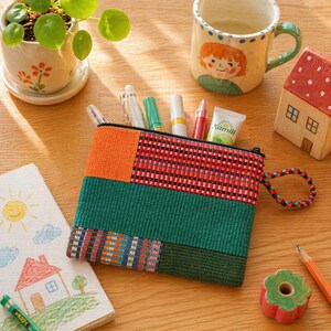 May include: A colorful, patchwork pencil case with a zipper, filled with pens and cosmetics. The case is orange, red, and green. A small ceramic house, a mug with a child's face, and a small potted plant are also on the wooden surface.