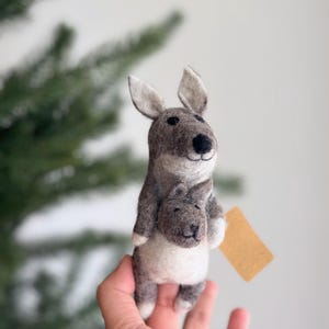 May include: A gray felt kangaroo holding a smaller gray felt joey in its pouch. The kangaroo is standing on a hand.