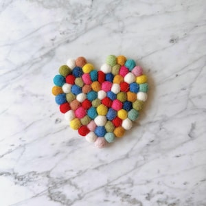 May include: A heart-shaped coaster made of colorful felt balls. The coaster is made up of small, round felt balls in a variety of colors, including red, blue, green, yellow, and white.