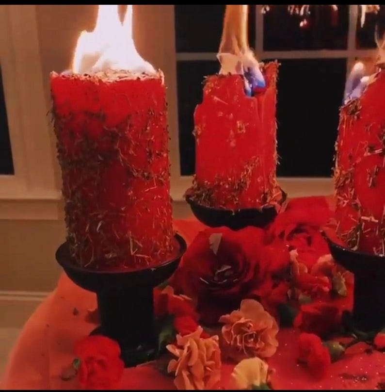 May include: Three red candles with flames burning, set on black candle holders. The candles are surrounded by red and pink flowers on a red surface.