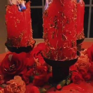 May include: Three red candles with dried herbs embedded in the wax are burning in black candle holders. The candles are surrounded by red and pink flowers.