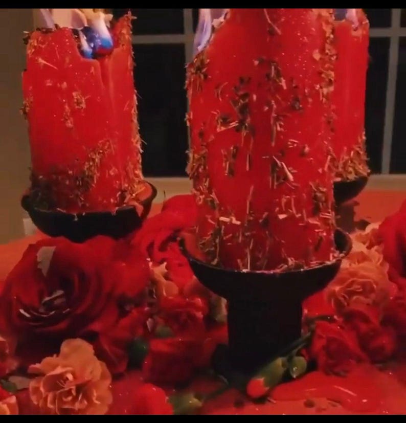 May include: Three red candles with dried herbs embedded in the wax are burning in black candle holders. The candles are surrounded by red and pink flowers.