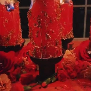 May include: Burning red pillar candles with dried herbs, set in black holders, surrounded by red roses and petals. The candles are lit, with wax dripping down. A spiritual or decorative arrangement.