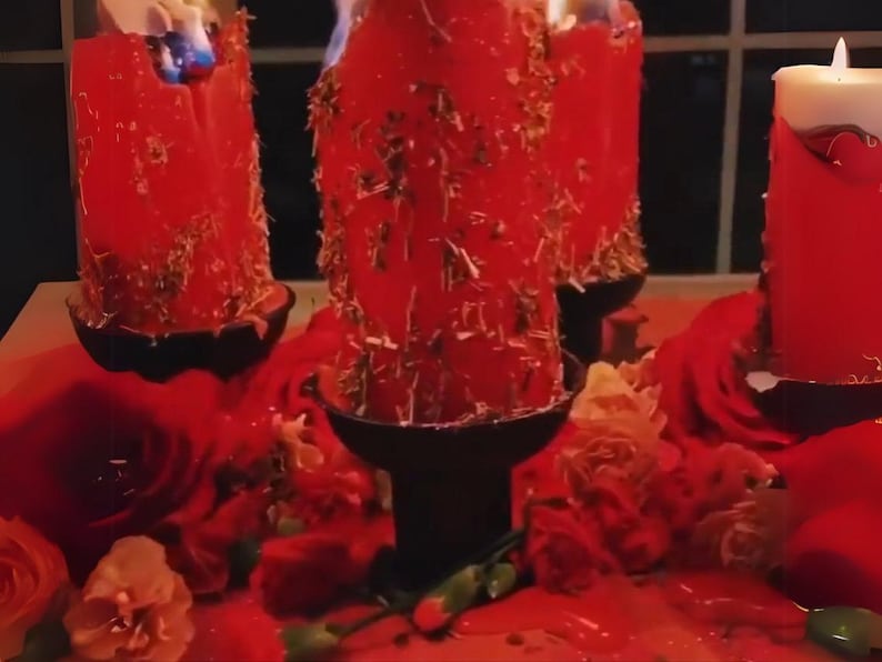 May include: Burning red pillar candles with dried herbs, set in black holders, surrounded by red roses and petals. The candles are lit, with wax dripping down. A spiritual or decorative arrangement.