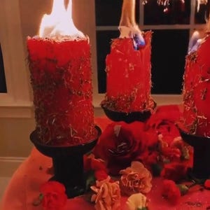 May include: Three burning red pillar candles with dried herbs are displayed on black candle holders. The candles are surrounded by red roses and other flowers. The scene is set on a red surface.
