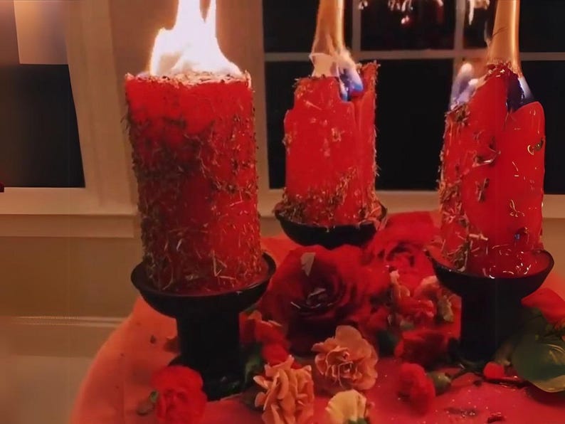 May include: Three burning red pillar candles with dried herbs are displayed on black candle holders. The candles are surrounded by red roses and other flowers. The scene is set on a red surface.