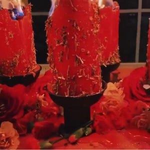 May include: Three red candles with a textured surface are burning on black candle holders. The candles are surrounded by red roses and other flowers.