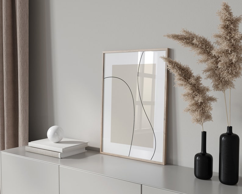 Peut inclure: Une impression abstraite encadr&eacute;e avec des lignes noires sur un fond beige est accroch&eacute;e &agrave; un mur blanc au-dessus d'une commode gris clair. Deux vases noirs avec de l'herbe de pampa s&eacute;ch&eacute;e sont sur la commode.