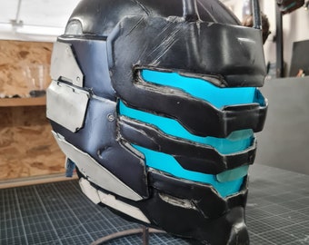 May include: A black and gray helmet with a futuristic design, featuring glowing blue accents. The helmet has a segmented appearance with silver-colored plates on the side. It is displayed on a metal stand, suggesting a collectible or cosplay item.