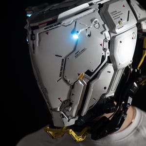 May include: A futuristic, silver and white helmet with intricate paneling and glowing blue lights. The helmet has yellow accents and black wiring. The text "PINCH POINT HAZARD" and "SLIDE TO OPEN" are visible.