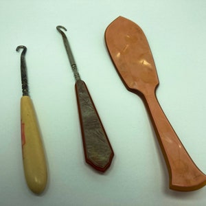 May include: Three vintage shoe horn tools. One is a long, thin, silver-colored tool with a wooden handle. The second is a short, curved, silver-colored tool with a wooden handle. The third is a long, flat, pink plastic tool.