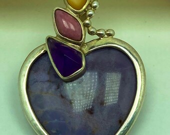 May include: A silver pendant with a heart-shaped purple stone. The pendant has a silver border and is adorned with three colorful gemstones: yellow, pink, and purple. The pendant is designed with a unique and artistic flair.