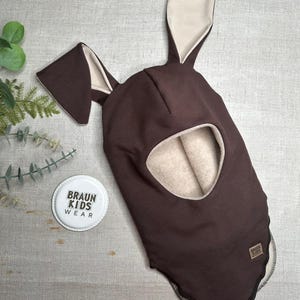 May include: A brown balaclava hat with long bunny ears and a face opening. The inside of the ears and face opening are a light beige colour. The hat has a tag that says "BRAUN KIDS WEAR".