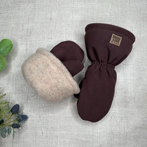 Può includere: Un paio di guanti da bambino marroni con un morbido polsino beige foderato in pile. I guanti hanno un'etichetta con la scritta "BRAUN KIDS WEAR". I guanti sono progettati per tenere calde le manine.