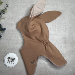 May include: A tan-colored balaclava-style hat with long, bunny-like ears. The hat has a light beige interior and a gathered section at the back. The hat has a small tag that says "BRAUN KIDS WEAR" and a round logo with the same text.