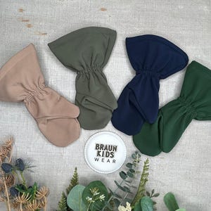 May include: Four baby mittens in different colors: tan, olive green, navy blue, and forest green. The mittens are made of soft fabric and have a gathered wrist. A white plate with the text "BRAUN KIDS WEAR" is also visible.