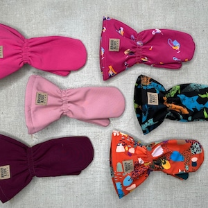 Puede incluir: Seis pares de mitones para niños en varios colores y estampados. Los colores lisos incluyen rosa, rosa claro y burdeos. Los mitones estampados presentan un diseño rosa oscuro, un estampado de dinosaurios y un diseño naranja. Cada mitón tiene una pequeña etiqueta que dice "BRAUN KIDS WEAR."