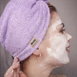 May include: A light purple terry cloth hair towel with a label that reads "BRAUN WEAR". The towel is wrapped around the head. The person in the image has a facial mask on.