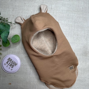 Könnte beinhalten: Eine hellbraune Sturmhaube mit hellbeigen Ohrakzenten. Die Mütze hat eine große Gesichtsöffnung und ein kleines Etikett mit der Aufschrift "BRAUN KIDS WEAR". Ein runder, violetter Knopf mit dem Text "BRAUN KIDS WEAR" ist ebenfalls sichtbar.