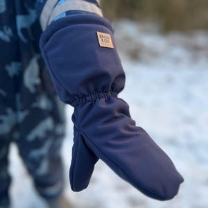May include: Navy blue mitten with a small brown label that reads "BRAUN KIDS WEAR". The mitten is part of a winter outfit. The background is blurred, suggesting an outdoor setting with snow.