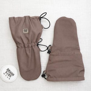 Op de afbeelding: Twee bruine kinderwanten met zwarte trekkoorden. De wanten hebben een klein vierkant label met de tekst "BRAUN KIDS WEAR". Een wit rond label met dezelfde tekst is ook zichtbaar.