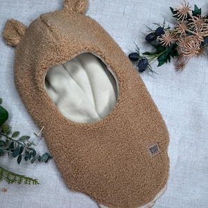 Könnte beinhalten: Eine hellbraune Sturmhaube mit zwei kleinen Bärenohren oben. Die Innenseite ist cremefarben. Der Hut hat eine weiche, flauschige Textur und ein kleines Etikett mit der Aufschrift "BRAUN KIDS".