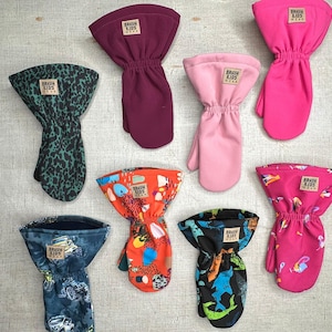 May include: A collection of children's mittens in various colors and patterns. The mittens include solid colors like pink and burgundy, and patterned designs with dinosaurs, trucks, and animal prints. Each mitten has a small label that reads "BRAUN KIDS WEAR."