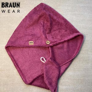 Könnte beinhalten: Ein magentafarbener Haartuch-Wrap mit Knopfverschluss und kleiner Schlaufe. Das Handtuch hat ein weiches, strukturiertes Aussehen und ein kleines Etikett mit dem Text "BRAUN WEAR".