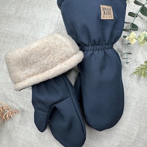 Puede incluir: Manoplas azul marino con puño de forro polar beige. Las manoplas tienen una muñeca elástica y una pequeña etiqueta marrón que dice "BRAUN KIDS WEAR". Diseñadas para niños.