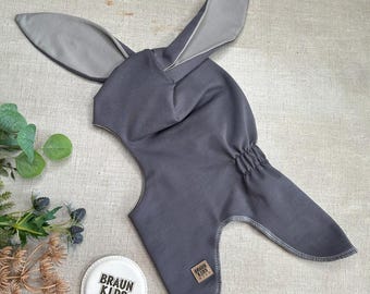 May include: A grey bunny hat with long, upright ears and a hood. The ears are lined with a lighter grey fabric. The hat has a drawstring at the back and a small label that says "BRAUN KIDS WEAR".