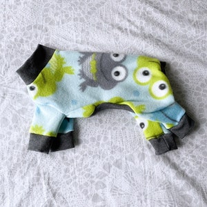 May include: A blue and green fleece dog pajamas with a monster print. The pajamas have grey trim around the neck, legs, and tail.