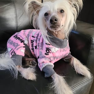 May include: A Chinese Crested dog wearing a pink and gray dog pajama set. The pajamas have a repeating pattern of white clouds and the words "I Love". The dog has white fur and is sitting on a black leather couch.