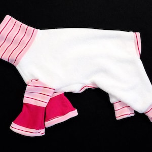 May include: White pet onesie with pink and red striped trim on the neck, sleeves, and legs. The legs are a solid bright pink color. The onesie is designed for a small dog or cat.