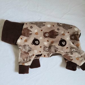May include: A beige pet onesie with a repeating pattern of brown bears and autumn leaves. The cuffs and neck are a solid brown color. The onesie is designed to cover the pet's body and legs.