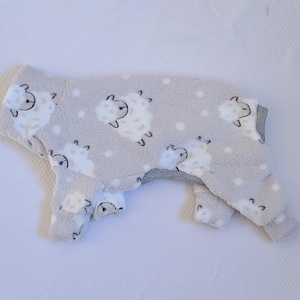 May include: A light gray onesie with a sheep pattern. The onesie is made of a soft, fuzzy material and features white sheep with black accents and small white dots. The garment has long sleeves and legs.