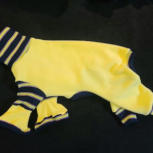 May include: Yellow dog jumpsuit with navy blue and yellow striped trim on the collar and leg cuffs. The dog outfit is made of a soft, plush material.