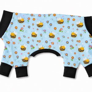 May include: A light blue pet bodysuit featuring a repeating pattern of yellow and black bumblebees, small orange and purple flowers, and tiny pink hearts. The garment has solid black cuffs at the four leg openings and the neck opening.