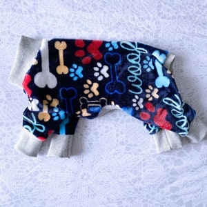 May include: A blue and gray dog onesie with a paw print and bone pattern. The onesie is made of soft fleece and has a comfortable fit.