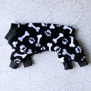 May include: Black and white dog pajamas with a bone and paw print pattern. The pajamas have black ribbed cuffs and a black collar.