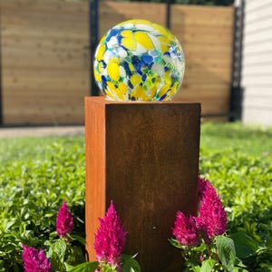 May include: A spherical glass garden ornament featuring a mottled pattern of bright yellow, deep blue, and white colors. The glass sphere is mounted on top of a tall, rectangular, weathered brown metal pedestal with a textured, rusted finish.