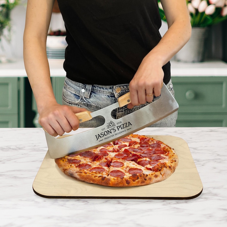 Puede incluir: Una persona usando un cortador de pizza para cortar una pizza de pepperoni en una tabla de cortar de madera. El cortador de pizza tiene un mango de madera y est&aacute; grabado con el texto "Jason's Pizza - Pizzeria d'Eccellenza - Est. 2008".