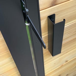May include: A matte black metal door latch mechanism mounted on horizontal light brown wooden planks. The hardware includes a vertical U-shaped bracket and a hinged arm attached to a black vertical frame, showing a functional locking system.