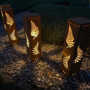May include: Several rectangular, brown metal garden lights with fern leaf cutouts. The lights are illuminated from within, casting a warm glow. They are placed on a bed of small stones, with greenery in the background.