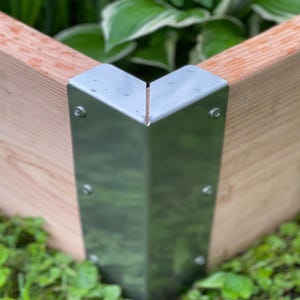 May include: Close-up of a corner of a wooden raised garden bed. The corner is reinforced with a silver metal bracket, secured with screws. The wood is light brown, and the metal has a reflective surface. Green plants are visible in the background.