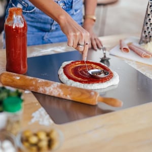 May include: A person is spreading tomato sauce on a pizza crust with a small ladle. A wooden rolling pin and a bottle of sauce are on the wooden surface. Sliced ham and cheese are in the background.