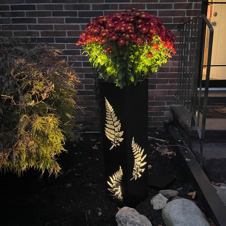 May include: A tall, black metal planter with illuminated fern leaf cutouts. The planter is filled with vibrant red and yellow mums. The design is lit from within, creating a warm glow. A stylish outdoor decor piece.
