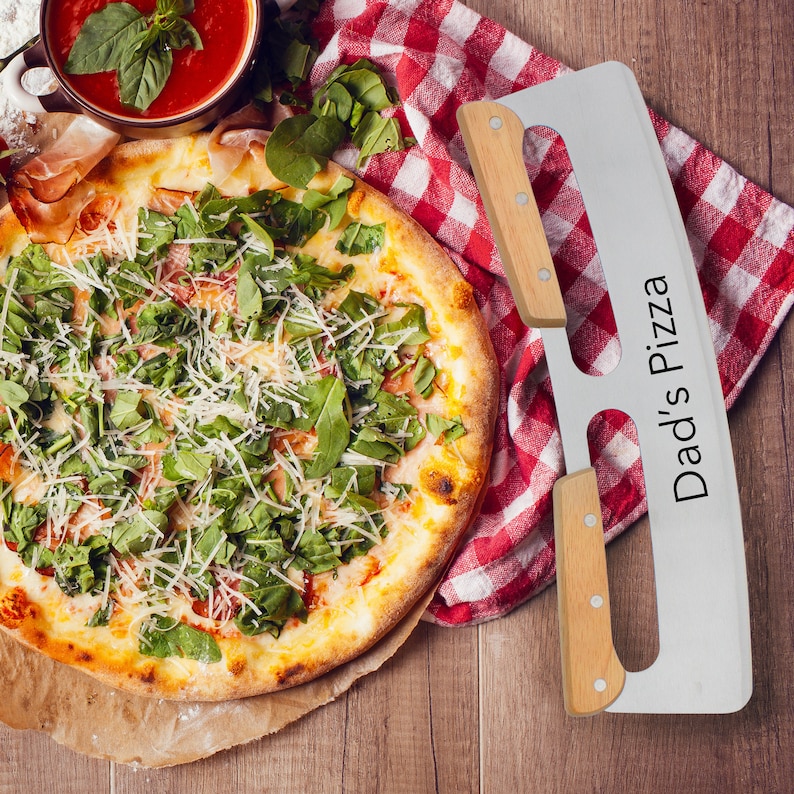 Puede incluir: Una pizza con r&uacute;cula, queso y jam&oacute;n sobre un fondo blanco. Un cortador de pizza de acero inoxidable con mango de madera est&aacute; al lado de la pizza. El cortador de pizza tiene las palabras "Dad's Pizza" impresas en &eacute;l.