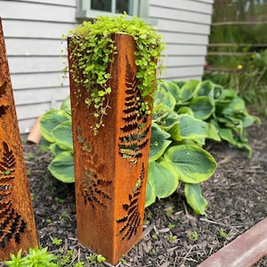 May include: A rectangular, weathered metal planter with fern cutouts and a vibrant green plant spilling over the top. The planter has a rusty orange hue and is set in a garden bed with lush greenery.